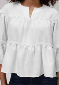 Hvit peplum-bluse med volanglag, utskjært hals og teksturert stoff. Ermene er vide og samlet nær mansjettene.