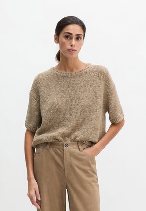 Γυναίκα με σκούρα μαλλιά μαζεμένα πίσω, φοράει μια φαρδιά μπεζ πλεκτή μπλούζα με κοντά μανίκια και ανοιχτοκαφέ παντελόνια ψηλόμεσα, με το ένα χέρι στην τσέπη.