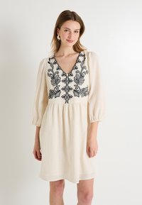 Vestido color crema con escote en V, mangas abullonadas y patrones florales bordados en negro. Fabricado con una tela texturada.