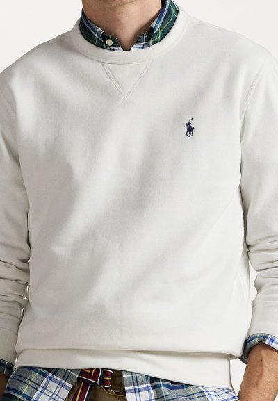 Man die een witte crewneck trui draagt over een blauw geruit overhemd met een klein marineblauw polo spelerlogo op de borst.