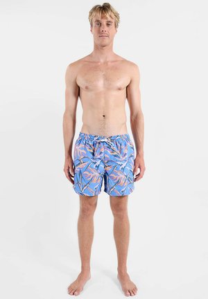 Homme blond debout pieds nus portant un short de bain bleu avec des motifs de feuilles orange et bleu foncé sur fond blanc.