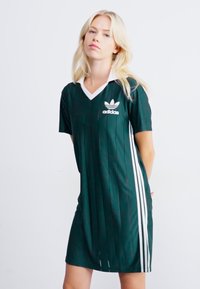 Zaļa Adidas kleita ar baltu apkakli un trim vertikālām strīpām pa sāniem. Izgatavota no teksturētas auduma ar smalkiem krokām.