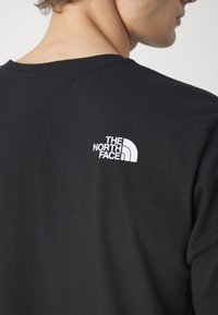 Mann trägt ein schwarzes T-Shirt mit weißem "The North Face"-Logo auf dem oberen Rücken, sichtbar von hinten und im Seitenprofil.