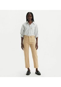 Camisa de botones a rayas blancas y marinas, con las mangas arremangadas, combinada con jeans de tiro alto color beige y mocasines negros, de pie contra un fondo neutro.