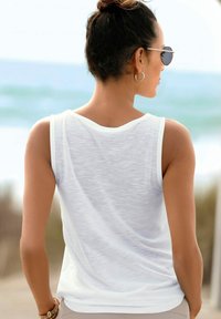 Top bianco senza maniche realizzato in tessuto leggero e dalla trama accentuata, con scollatura rotonda, indossato con pantaloni beige e un bracciale.