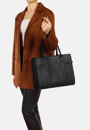 Borsa a tracolla nera in pelle con finitura testurizzata, dotata di doppi manici e logo embossato. Indossata con una giacca in suede marrone e pantaloni neri.