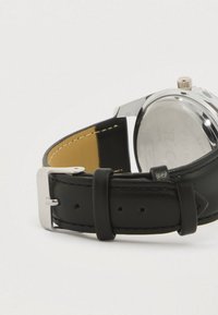 Λουρί ρολογιού από μαύρο δέρμα με ασημένια μεταλλική αγκράφα, με λεία υφή και αντίθετο ανοιχτό εσωτερικό. Ο ατσάλινος μηχανισμός είναι ορατός στο πίσω μέρος.