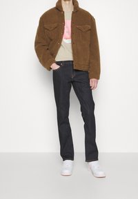 Homme portant une veste en polaire marron, un t-shirt graphique beige, un jean bleu foncé et des baskets blanches, se tenant avec une main dans la poche.