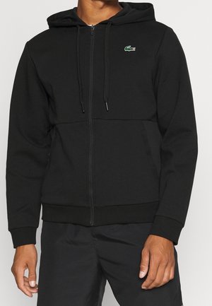 Mann trägt einen schwarzen Zip-up-Hoodie mit Kapuze und kleinem grünen Krokodil-Logo auf der Brust, kombiniert mit schwarzen Hosen.