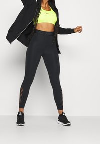 Femme portant un brassard de sport jaune néon, une veste noire ouverte, des leggings noirs taille haute avec des détails ajourés, et des chaussures de sport noires.