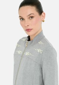Giacca grigia con zip, caratterizzata da un tessuto testurizzato, colletto alto e dettagli decorativi in perle sulle spalle. Dettaglio della zip dorata in evidenza.