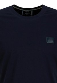 Navyblauwe katoenen t-shirt met een ronde halslijn, voorzien van een gestructureerde donkerblauwe patch met logo op de borst. Plat gestikte naden, korte mouwen.