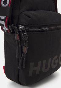 Borsa a tracolla nera in nylon con tasca frontale con zip, dettagli testurizzati e un audace logo "HUGO" in finitura opaca. Inclusa tracolla regolabile.