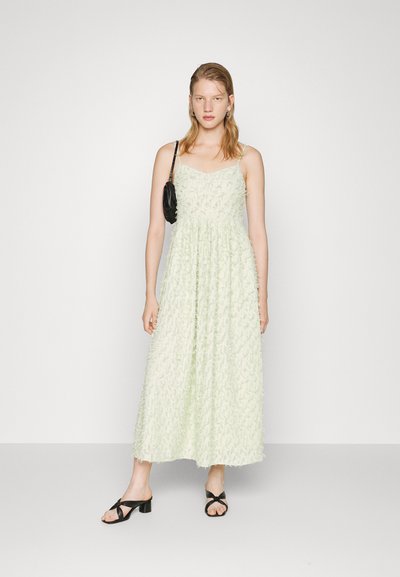 Vestido maxi verde claro com tecido texturizado, alças finas e forma evasê. Estilizado com uma pequena mala preta e sandálias de salto pretas.
