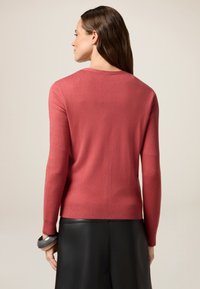 Pehmeä, korallinvärinen neule, jossa on pyöreä kaula-aukko, pitkät hihat ja ribbikuviointi hihoissa ja helmassa, yhdistetty mustan nahkahameen kanssa.