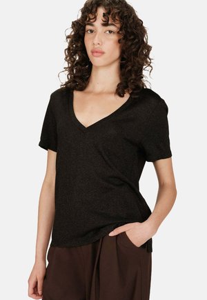 Une femme aux cheveux bouclés et foncés portant un haut noir à encolure en V et des pantalons marron, se tenant avec une main dans la poche contre un fond blanc.