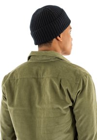 Marineblå ribbet strik beanie og olivengrøn corduroy skjorte, set fra baghuset. Skjorten har en button-down krave og en diskret tekstur.