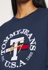 Granatowa bluza z okrągłym dekoltem, zdobiona wyszywanym logo "Tommy Jeans" oraz napisem "USA 1985" w białych i złotych akcentach.