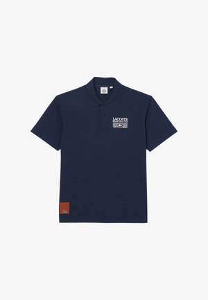 Polo Lacoste bleu marine avec logo du club Roland Garros et graphisme de filet de tennis sur la poitrine, petite pièce rouge avec logo crocodile près de l'ourlet.