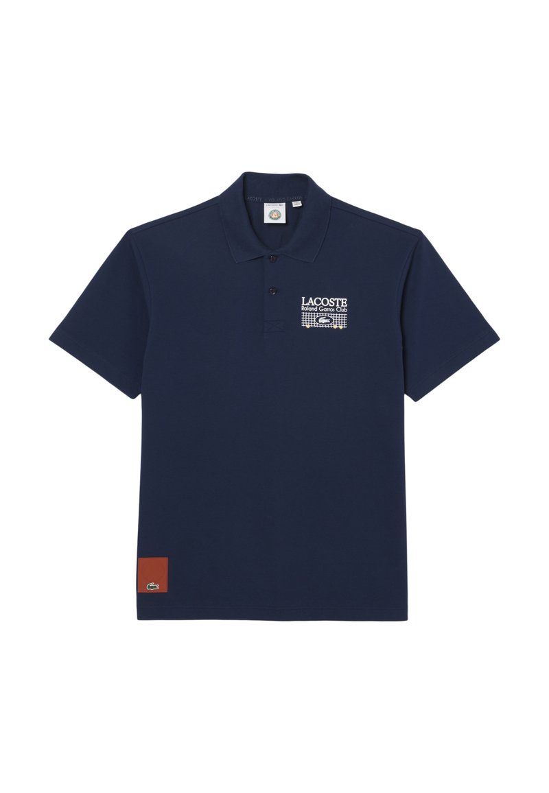 Polo Lacoste bleu marine avec logo du club Roland Garros et graphisme de filet de tennis sur la poitrine, petite pièce rouge avec logo crocodile près de l'ourlet.