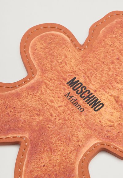Acessório castanho semelhante a couro em forma de um ursinho de pelúcia com uma superfície texturada. Apresenta costura branca e texto impresso "MOSCHINO Milano".