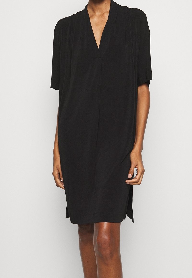 Svart kortärmad klänning med V-ringning, tillverkad av mjukt tyg, med subtila veckdetaljer och avslappnad passform, som når till mitten av låret.