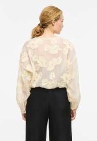 Blouse crème fluide avec broderie florale, texture transparente et poignets froncés, portée sur un pantalon noir taille haute. Vue de dos.