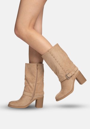 Piernas con botas de gamuza color beige hasta la mitad de la pantorrilla, con tacones de bloque apilados, cremallera lateral y detalle decorativo de hebilla sobre un fondo liso.