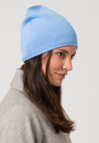 Berretto lavorato a maglia di colore azzurro chiaro con un top arrotondato, texture liscia e risvolto, indossato sopra capelli castano scuro di lunghezza media.