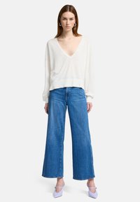 Maglione bianco a scollo a V, morbido e ampio, con maniche lunghe, abbinato a jeans blu a gamba larga e scarpe punta leggera di colore viola chiaro.