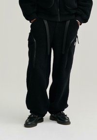 Czarne dresy z polaru z zamykanymi kieszeniami, regulowanymi paskami i luźnym krojem. Materiał ma fakturowany, miękki wygląd.