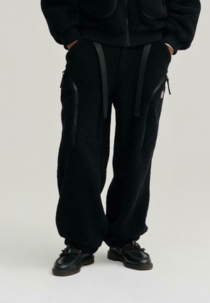 Pantalones de chándal de lana negra con bolsillos con cremallera, tirantes ajustables y un corte relajado. La tela tiene un aspecto suave y texturizado.