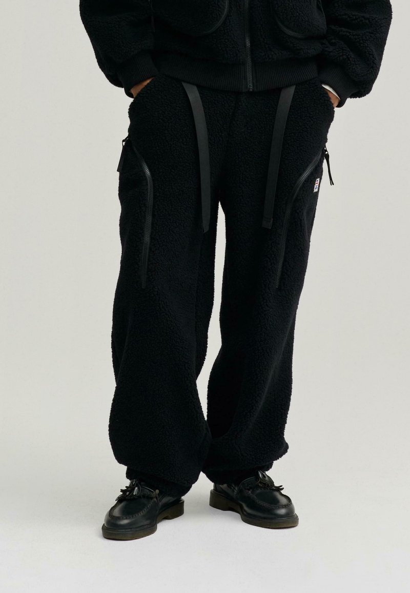 Czarne dresy z polaru z zamykanymi kieszeniami, regulowanymi paskami i luźnym krojem. Materiał ma fakturowany, miękki wygląd.