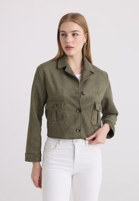 Femme portant une veste boutonnée vert olive avec poches à rabat, pantalon blanc, regardant légèrement sur le côté sur un fond clair uni.