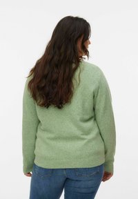 Femme aux longs cheveux bruns portant un pull vert clair et un jean bleu, vue de dos sur un fond blanc uni.