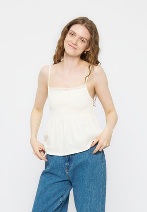 Jeune femme aux cheveux ondulés portant un haut blanc sans manches avec bordure en dentelle et un jean bleu large, se tenant devant un fond uni.