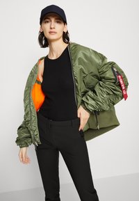 Olive green bomber jacket with an orange inner lining, ribbed cuffs, and zipper details, paired with a black tank top and black pants.