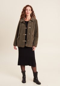Olivgrüne Fleecejacke mit Kragen, vier Knöpfen und zwei Vorderseiten-Taschen, kombiniert mit einem schwarzen Kleid und kniehohen schwarzen Stiefeln.