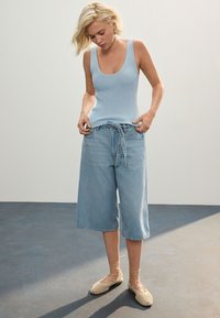 Svetlo modra rebrasta majica brez rokavov, ohlapne svetlo denim culottes hlače in bež čipkaste balerinke, postavljene na mehko, nevtralno ozadje.