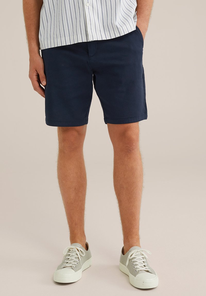 Muž v námořnických šortkách, světle šedých teniskách s bílou podrážkou a zapnuté pruhované košili, stojí se založenýma rukama v kapsách.