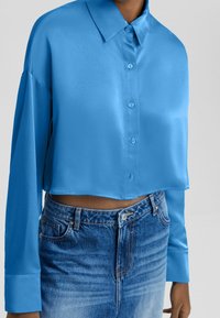 Donna che indossa una camicia satinata blu brillante, a maniche lunghe e con bottoni, abbinata a jeans a vita alta, sbiaditi.