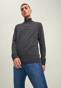 Sudadera gris oscuro con cremallera y cuello alto, puños ajustados y tejido texturizado. Combinada con jeans azules en una pose casual de pie.