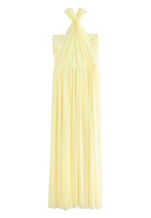 Nainen pukeutuneena vaaleankeltaiseen hihattomaan mekkoon, jossa on taakse sidottu rusetti, katsoo olkansa yli yksiväristä taustaa vasten.