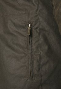 Dark green jacket fabric with a smooth texture, featuring a zippered side pocket and visible stitching along seams.