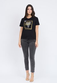 Camiseta de algodón negra con un estampado gráfico en dorado y plateado, cuello redondo, mangas cortas, combinada con jeans ajustados grises y sandalias de tacón doradas.