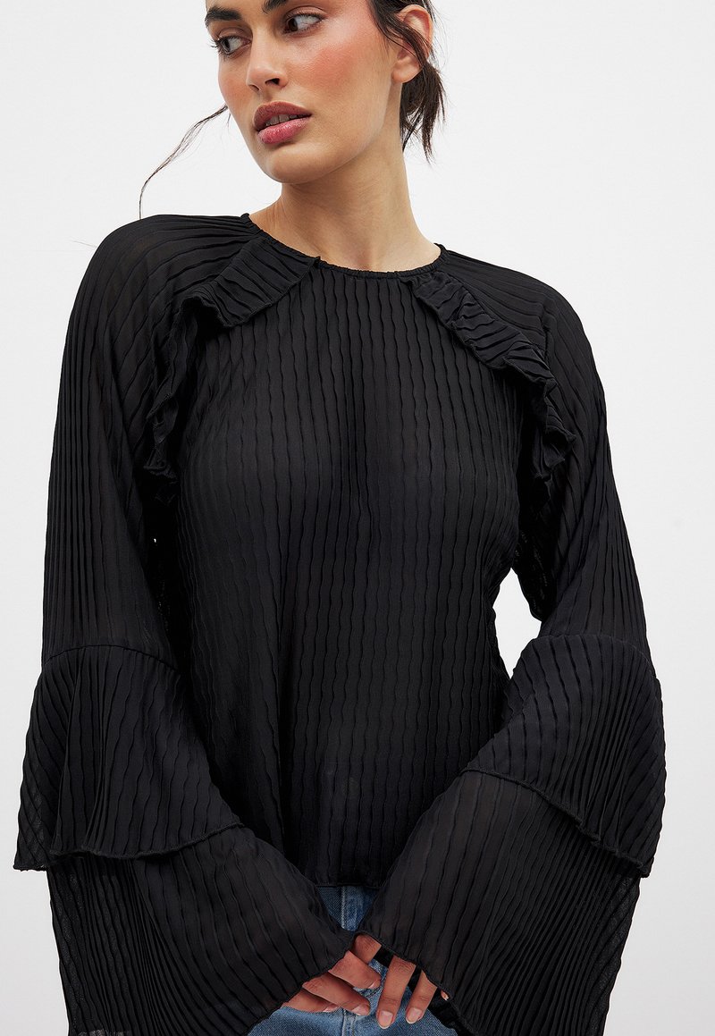 Black pleated blouse with long, flared sleeves and ruffled neckline. Made of lightweight fabric, featuring a smooth texture and subtle sheen.