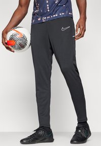 Schwarze Nike Sporthose mit schmalem Schnitt, Seitentaschen und einem weißen Logo. Das Modell hält einen schwarz-weiß gemusterten Fußball.