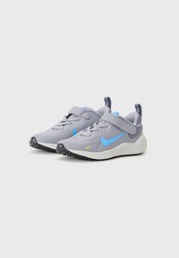 Chaussures de sport grises avec une tige en maille breathable, accents bleus, sangle Velcro et semelle extérieure noire texturée. Présente un détail de logo jaune.