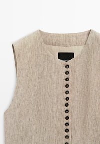 Mouwloos beige linnen top met een ronde halslijn en een verticale rij zwarte knopen aan de voorkant.