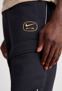 Czarne dresy Nike z złotym owalnym logo z napisem "AIR", z teksturowaną łatką i białymi akcentami szycia w pobliżu. Zbliżenie dłoni opierającej się na spodniach.